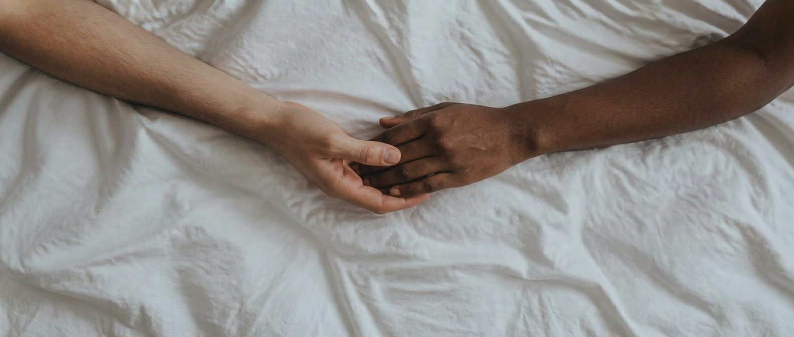 An interracial couple's hands resting together on white bed sheets